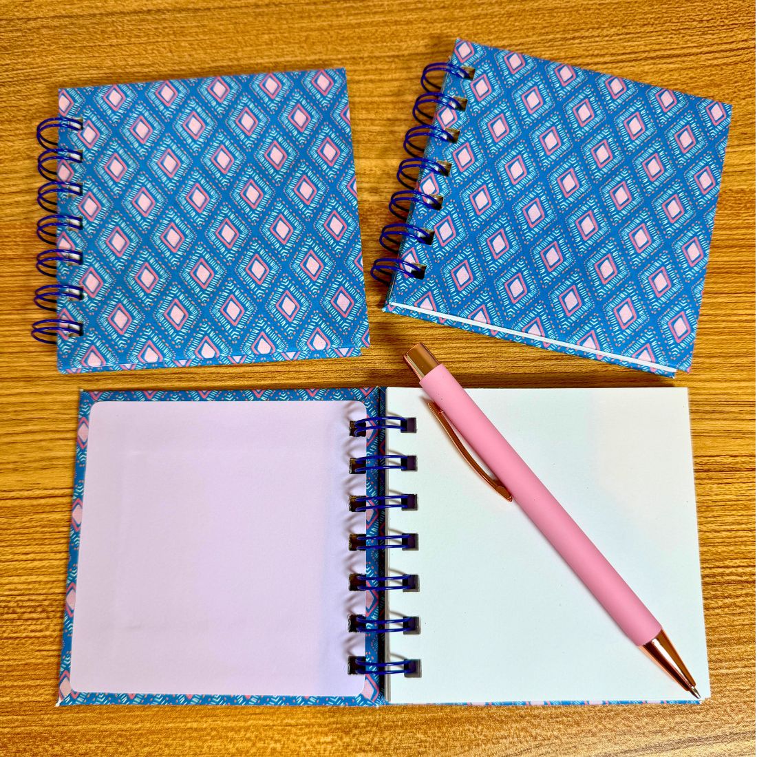 Two spiral-bound notebooks with a blue geometric pattern on a wooden surface, one open with a pink pen.