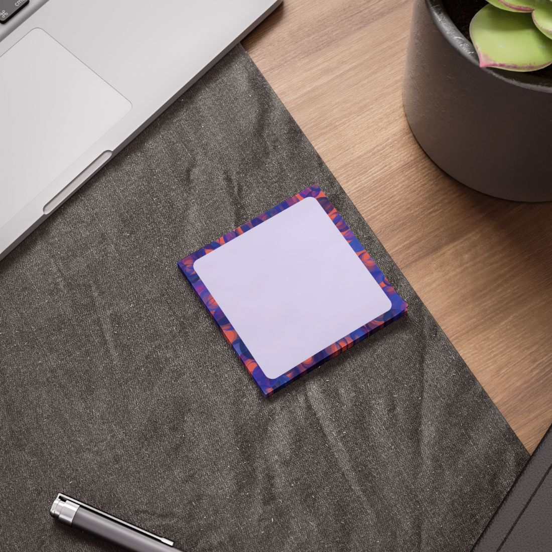 A square sticky note pad with a colorful patterned border and a blank white center, placed on a gray desk mat on a wooden desk next to a laptop, pen, and plant pot.