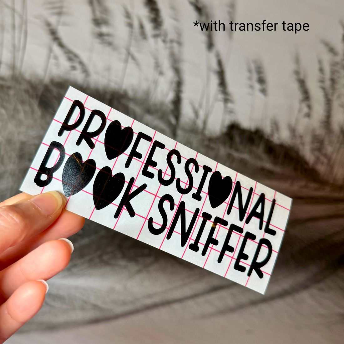 A hand holding the Professional Book Sniffer decal featuring heart accents and a red grid transfer tape overlay.