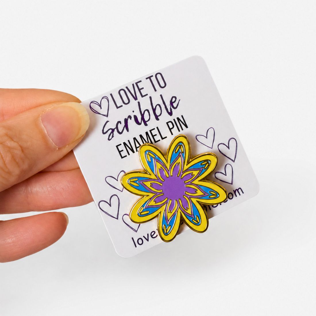 A hand holding a white display card featuring a colorful flower-shaped enamel pin. The pin has yellow outer petals, blue inner details, and a purple center.