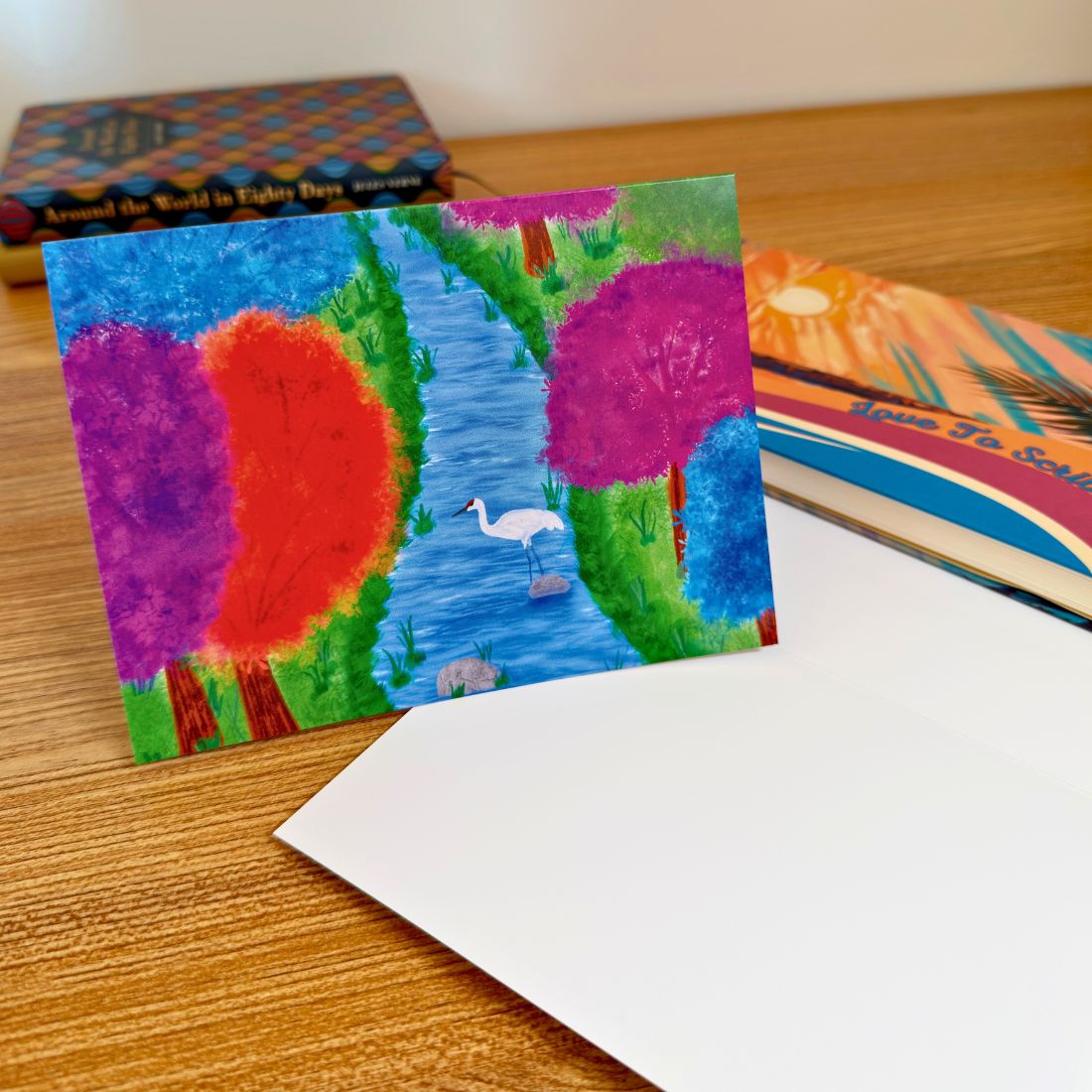 Love To Scribble greeting card showing a white crane in a blue river surrounded by vibrant purple, pink, orange, and blue forest trees on a wooden desk