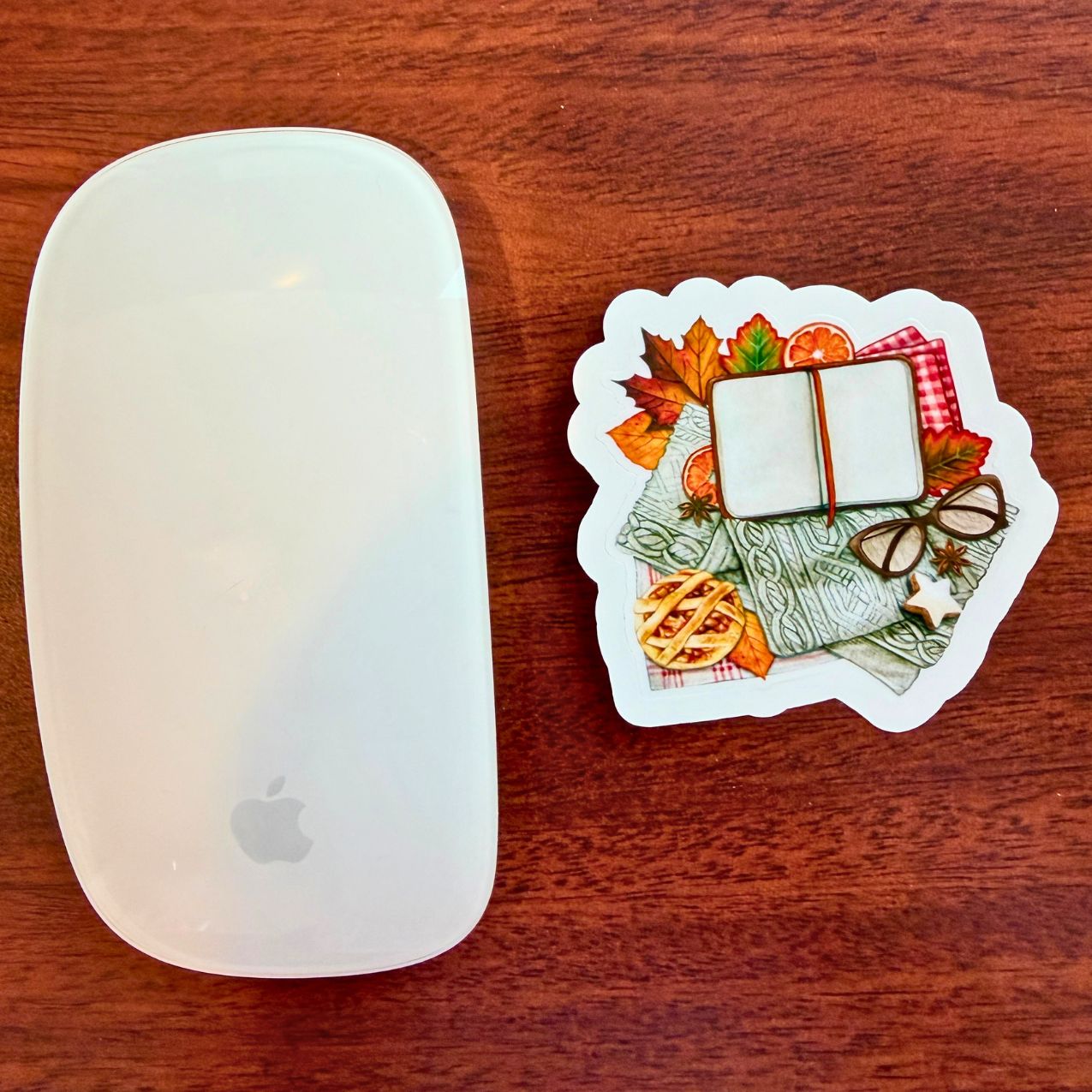 A white mouse is shown next to a die-cut sticker featuring an autumn-themed book illustration, placed on a dark wood surface.