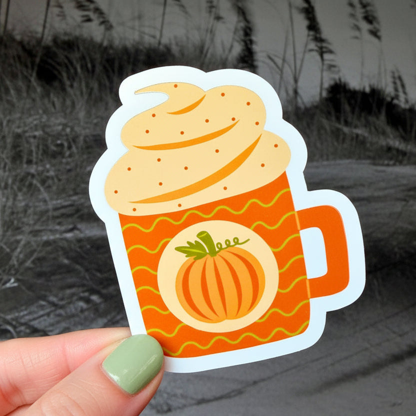 A hand holding a die-cut sticker shaped like a frothy orange mug with a pumpkin graphic, set against a blurred black-and-white beach scene.