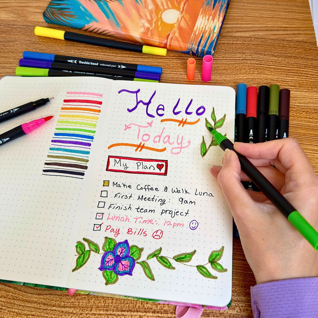 A hand holding a green-tipped marker writes in a decorated bullet journal page on a wooden desk, surrounded by colorful pens and notebooks.