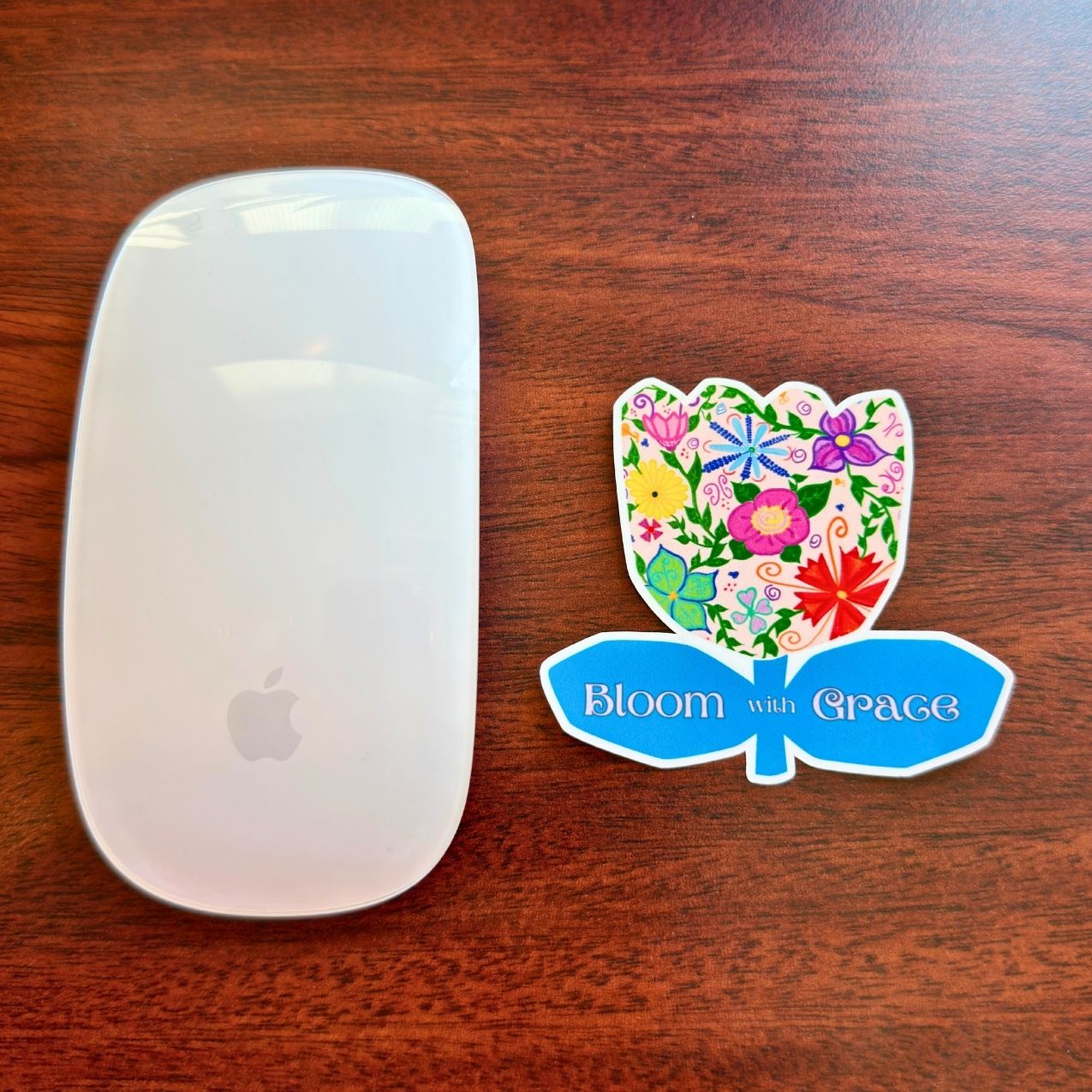 A white Apple Magic Mouse is placed on a dark brown wooden table next to a colorful flower-shaped sticker that reads 'Bloom with Grace'.