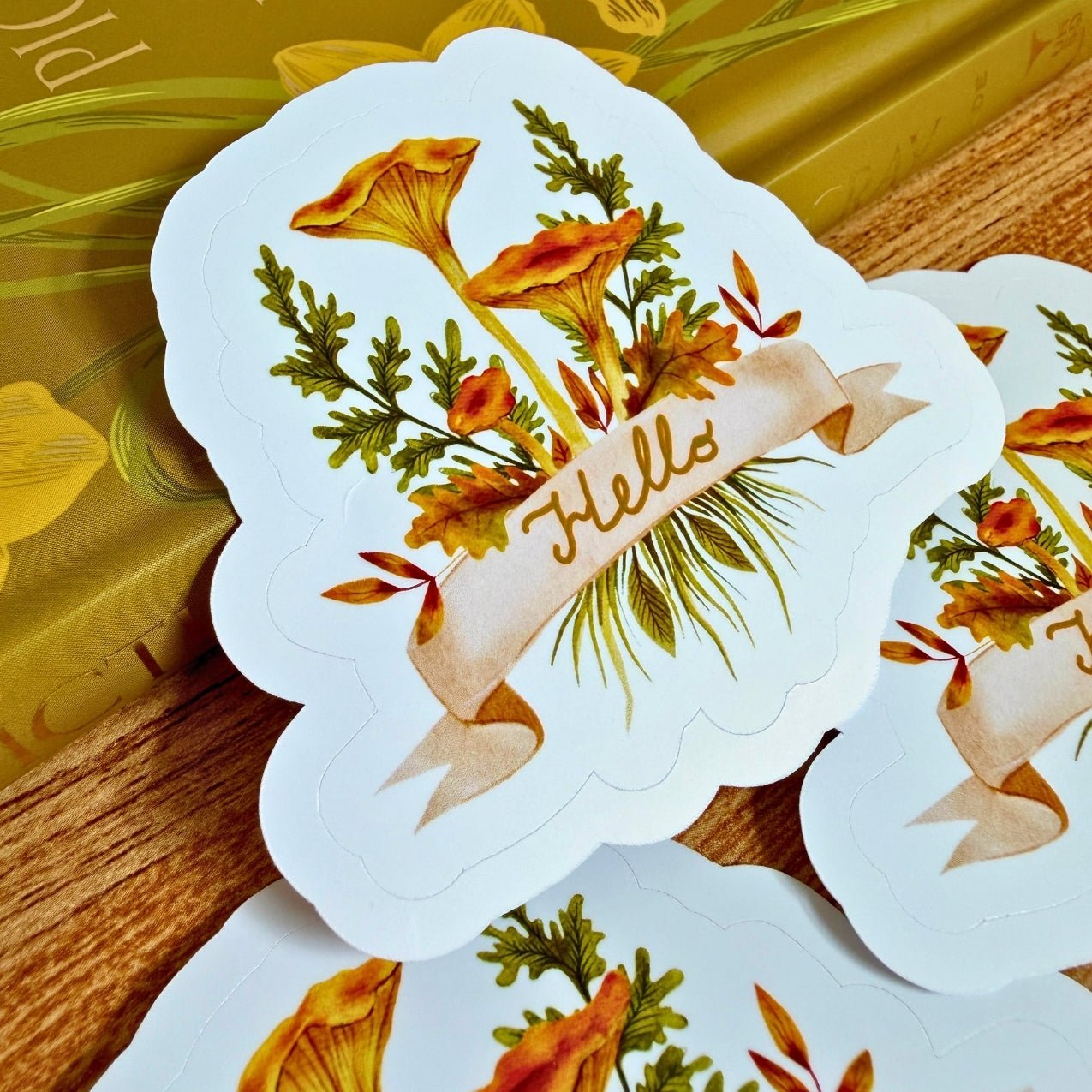 Love To Scribble white sticker featuring an autumn floral illustration with yellow-orange flowers, green leaves, and a beige ribbon banner that reads 'Hello,' shown on a wooden surface with additional matching stickers partially visible.