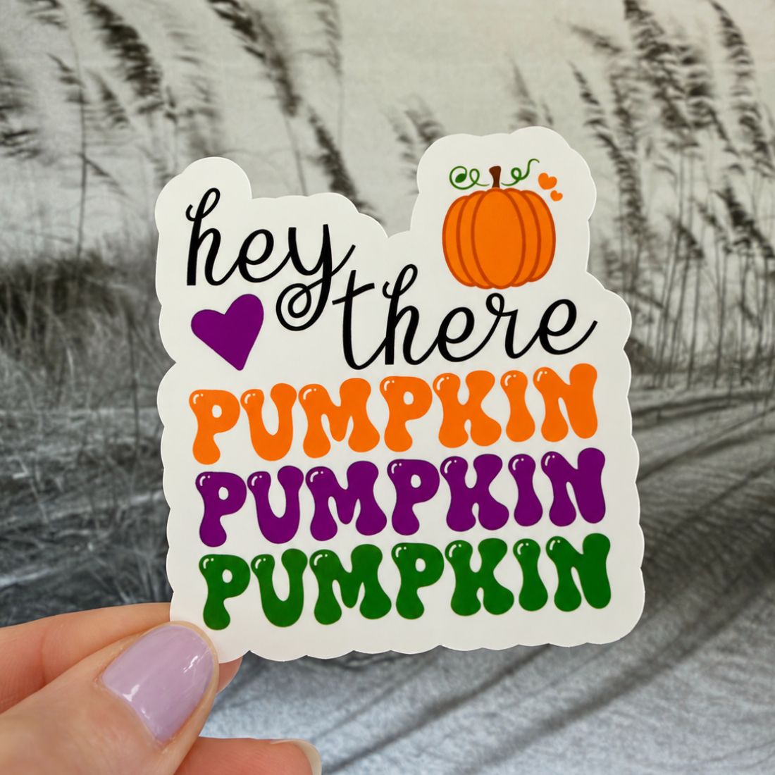 A hand with lavender nail polish holds a die-cut sticker that reads 'hey there PUMPKIN PUMPKIN PUMPKIN' in black, orange, purple, and green lettering, with a small orange pumpkin illustration. The background is a soft-focus gray and white patterned fabric.