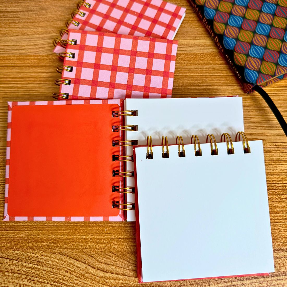 Multiple handmade pink and orange square mini notebooks stacked and styled open on a wood surface to show the blank interior and lay-flat binding.