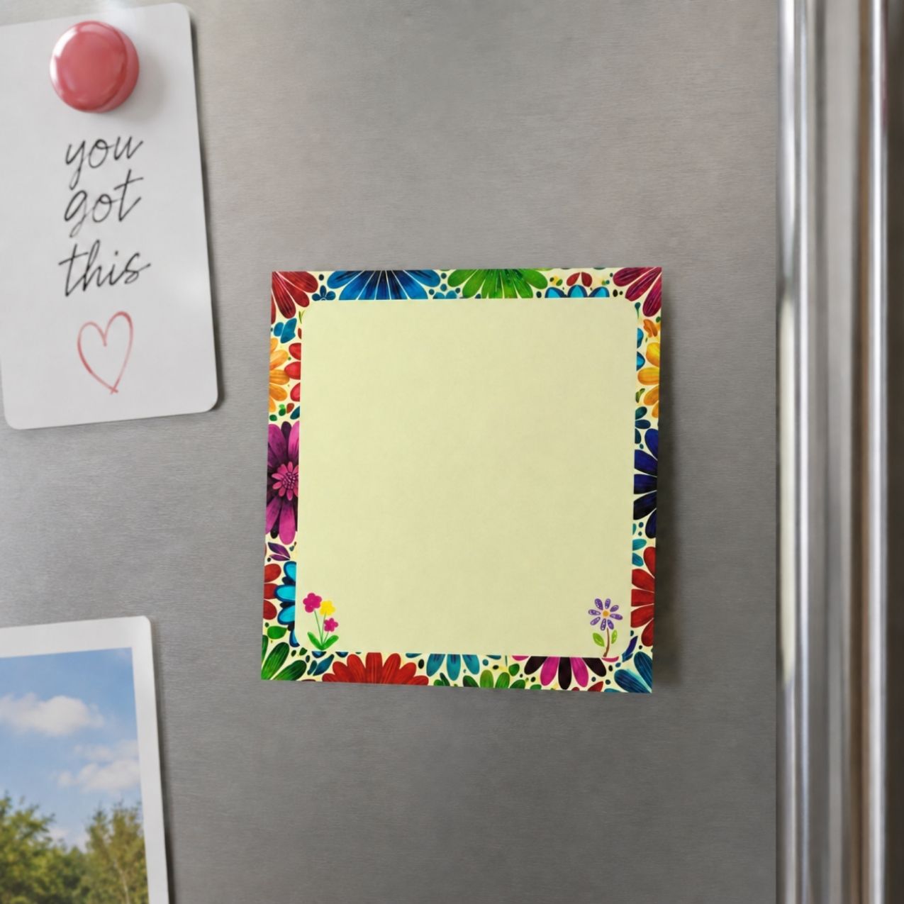 A square yellow sticky note pad with a colorful floral border is attached to a stainless steel refrigerator. A white note with a pink magnet and a partial photo are also visible on the fridge.
