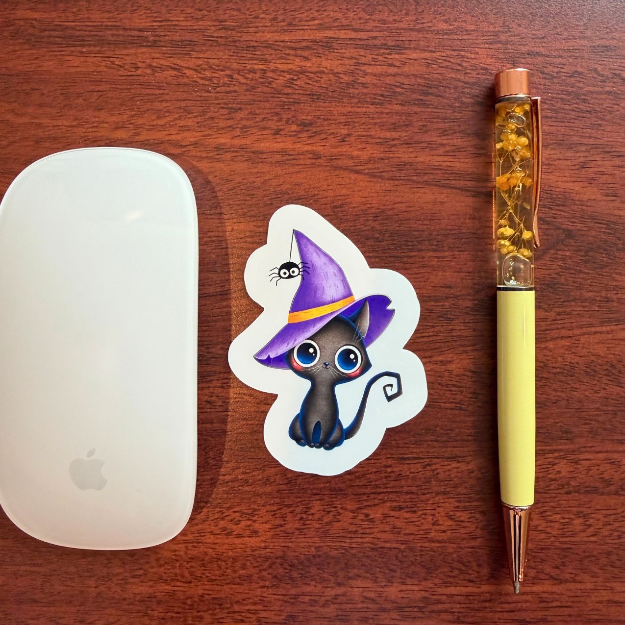 A die-cut sticker featuring a cute black cat wearing a purple witch hat with a yellow band, displayed on a dark wood tabletop next to a white computer mouse and a yellow pen.