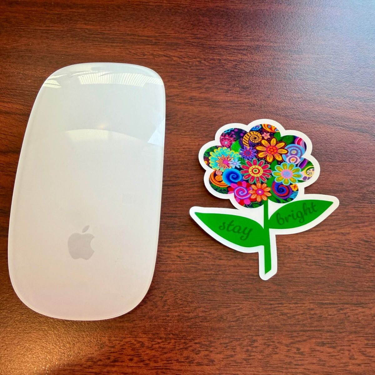 A white Apple Magic Mouse next to a colorful die-cut sticker shaped like a flower with the words 'stay bright' on a dark wood tabletop.