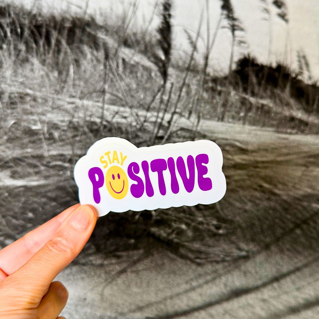 A hand holds a die-cut sticker that reads 'STAY POSITIVE' in purple and yellow text with a smiley face, set against a blurred black-and-white outdoor background.
