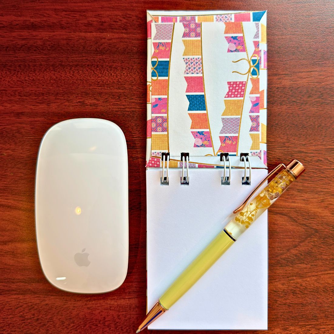 Top-down view of an open White Extra Mini Notebook revealing its smooth blank pages, styled on a dark wood desk next to a white computer mouse and a yellow floral pen.